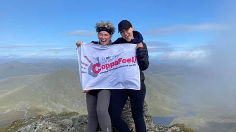 Photo showing Maxine Laceby and Emma Willis, two white women wearing dark hiking clothes, standing on a mountain against a cloudy blue sky and holding up a banner which says "Coppafeel!"