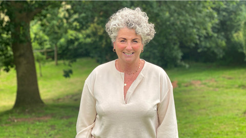 Photo showing Maxine Laceby, a white woman in her fifties with short silver hair, she is wearing a beige top and smiling at the camera against a background of grass and trees