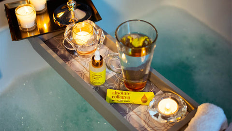 Photo showing a relaxing bubble bath with a wooden bath tray that contains candles, a latte glass, Absolute Collagen sachet and Maxerum bottle