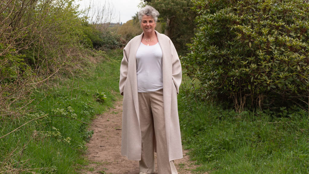 Photo showing Maxine Laceby, a white lady with short curly silver hair, standing outside by some hedges and wearing a long cream coat