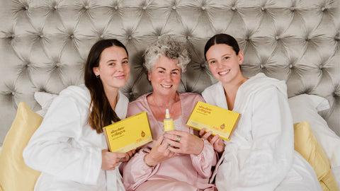 Photo showing Maxine, Margot and Darcy Laceby smiling at the camera, they are sitting against a grey velvet headboard and wearing dressing gowns and they are holding up Maxerum bottles and Absolute Collagen boxes