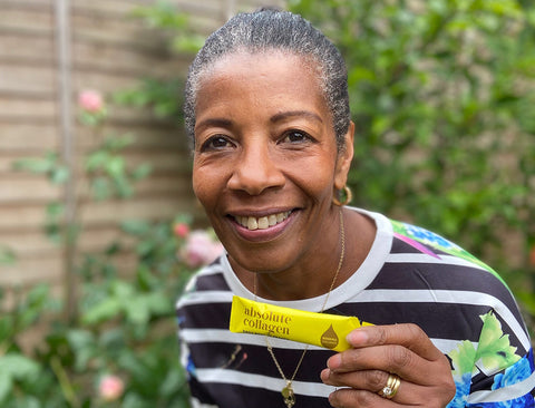 Photo of a smiling Black woman with short hair and a striped t-shirt, she is holding up a yellow Absolute Collagen sachet