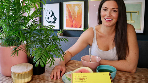 Photo showing a smiling white woman with long brunette hair sitting at a table with food and a yellow box of Absolute Collagen
