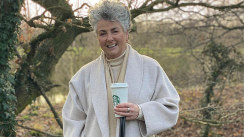 Photo showing Maxine Laceby standing outside by a tree, she is smiling and wearing a long cream coat and holding a reusable coffee cup, and her lurcher Rosie is standing by her