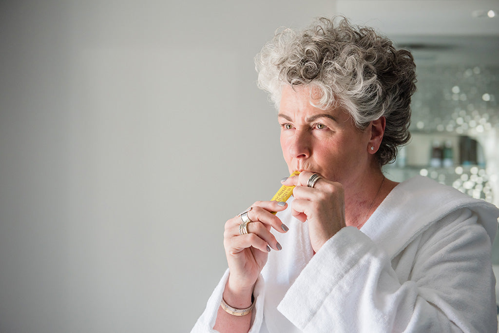 Photo of a white woman with short silver hair wearing a white dressing gown and squeezing a yellow Absolute Collagen sachet into her mouth
