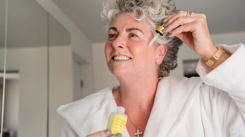 Photo showing Maxine Laceby wearing a white dressing gown and applying Maxerum to her face while smiling into a mirror