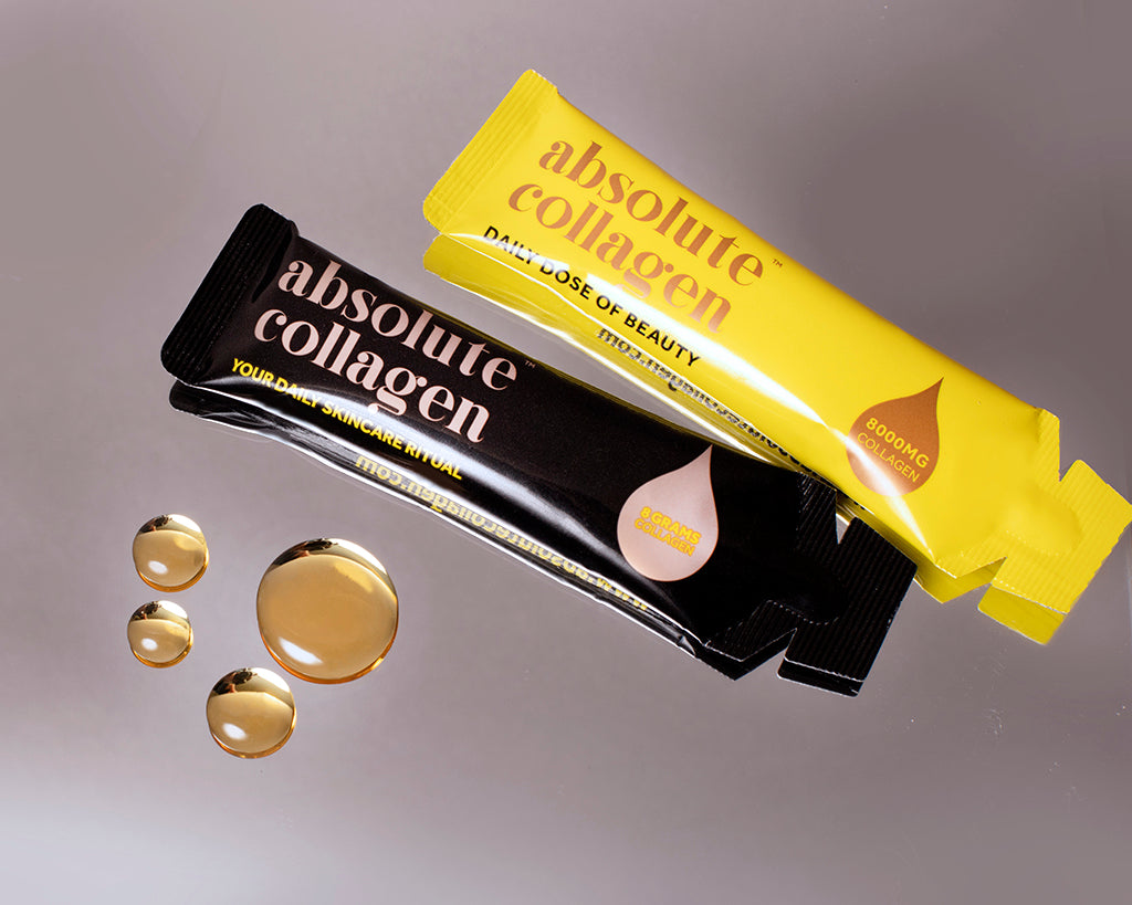 Photo of two sachets of Absolute Collagen, one yellow sachet and one black, lying on a grey surface, with droplets of liquid collagen next to them. 