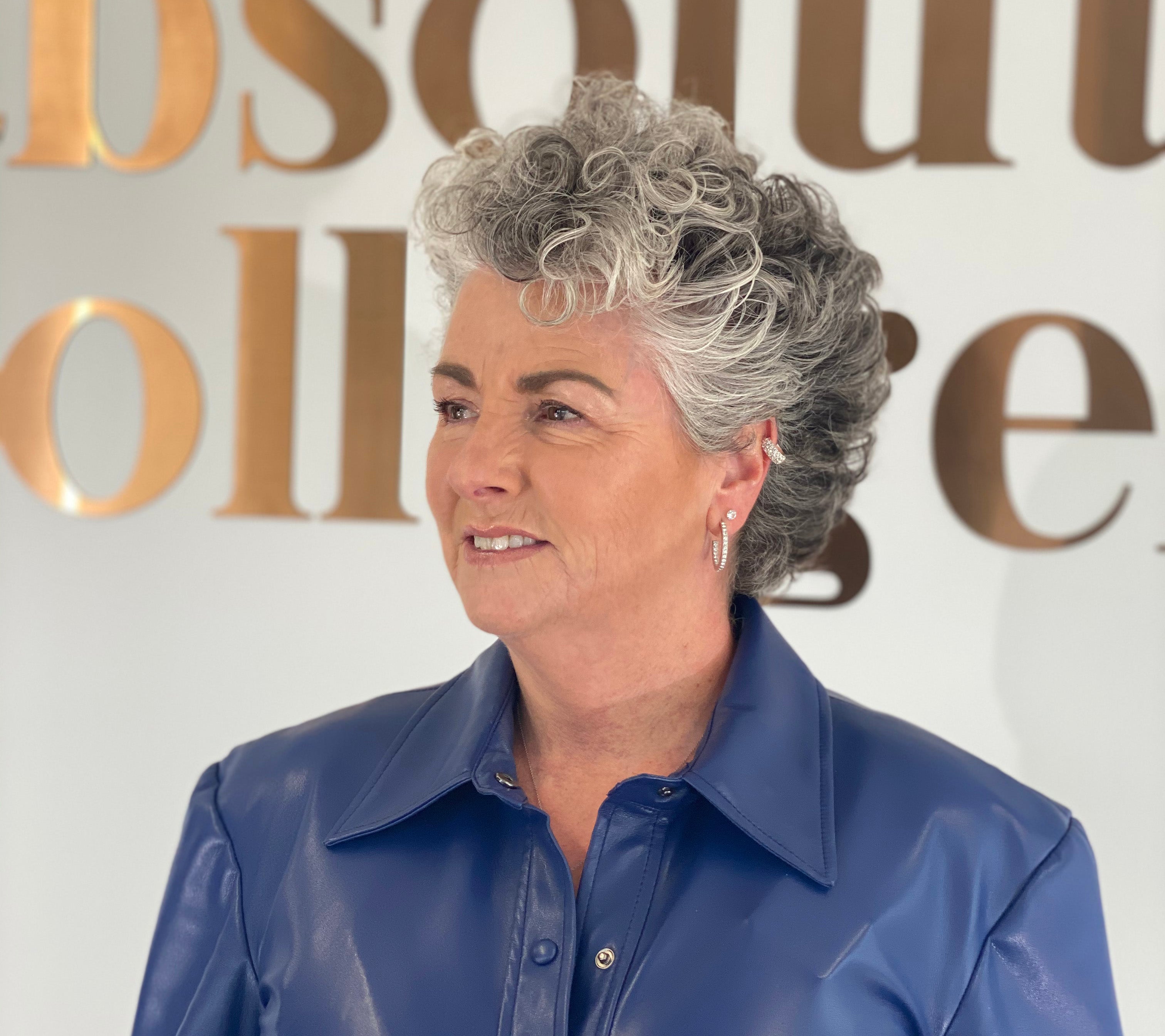 Photo of a white woman with short silver wavy hair and a blue leather top, she is looking to the left and smiling slightly